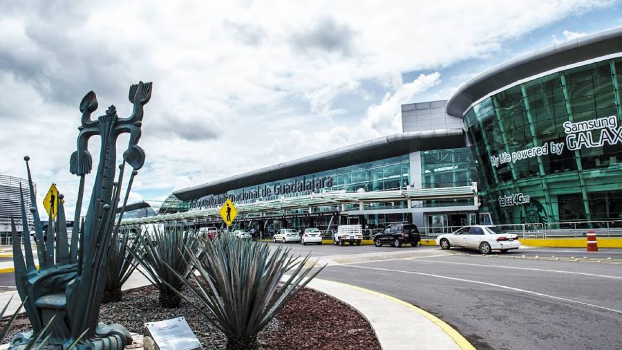 Fachada principal del Aeropuerto Internacional de Guadalajara, con su moderna estructura de vidrio y metal. En primer plano, una escultura representativa de la región junto a plantas de agave, símbolo de la identidad jalisciense. Vehículos circulan en la zona de arribos y salidas, mientras el cielo nublado enmarca la escena.