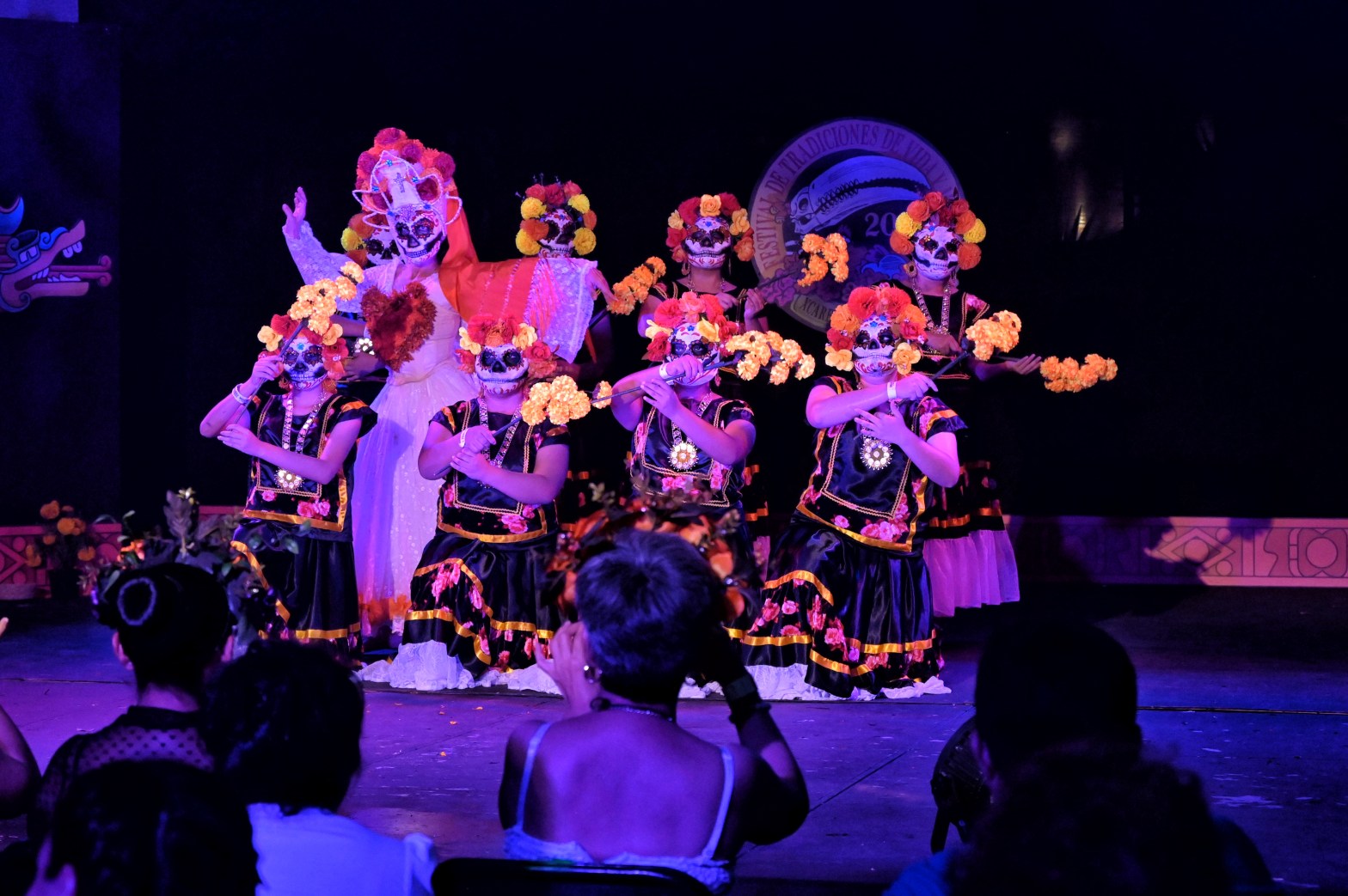 Grupo de bailarinas ataviadas con trajes tradicionales y maquillaje de calaveras, realizando una coreografía durante una celebración cultural en Michoacán, iluminadas por luces moradas y rodeadas de flores de cempasúchil. Ellas muestran la fuerza visual y simbólica de las tradiciones michoacanas como parte esencial del patrimonio cultural vivo del estado.