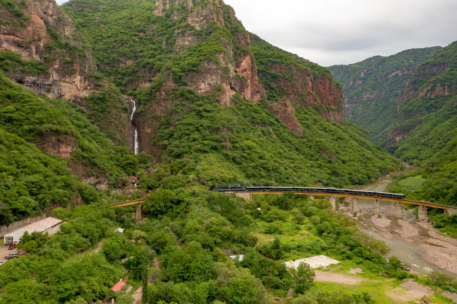 El tren Chepe atraviesa un puente en las Barrancas del Cobre, rodeado por montañas cubiertas de vegetación y un cañón atravesado por un río en el estado de Chihuahua